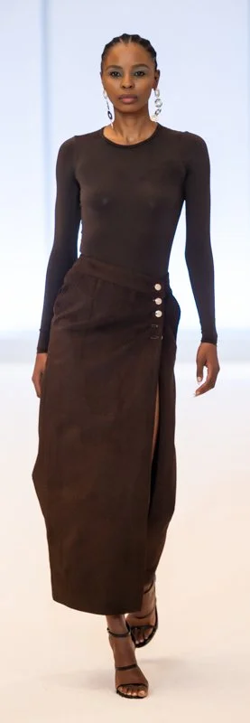 Model walking on runway wearing a long-sleeved brown top and high-slit brown skirt with button details, accessorized with earrings and black strappy heels.