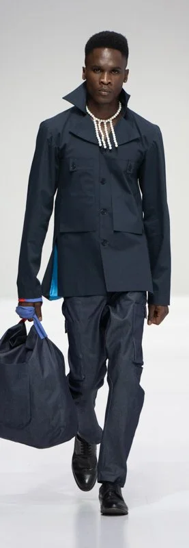 Model walking on runway in dark blue outfit with high collar, carrying a large matching bag