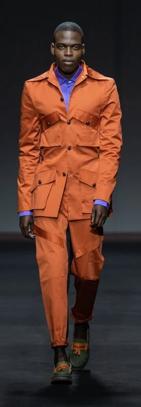A male model walking on a runway wearing an orange suit with a blue shirt and stylish green shoes with orange accents.