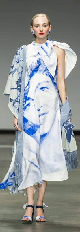 Model walking down runway wearing a high-fashion outfit with a large face print on a long dress, paired with a fringed blue and white shawl, and blue strappy heels.