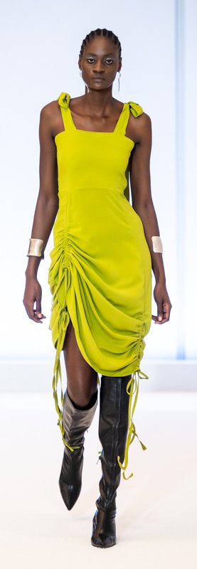 Model wearing a bright yellow dress with ruched design, black knee-high boots, and silver bracelets on a runway.