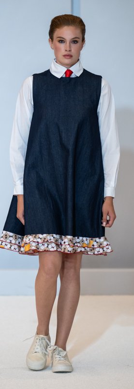 A person wearing a denim dress over a white shirt with a red tie, standing on a light-colored surface. The dress has a floral trim at the hem, and the person is wearing white sneakers.