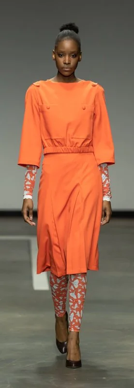 Model walking on a runway wearing an orange dress with patterned leggings and dark shoes.