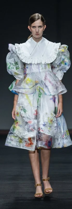 Model wearing a colorful, abstract-patterned dress with puffed sleeves and ruffled collar on a runway.