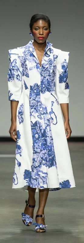 Model walking runway in white and blue floral dress with bold shoulders.