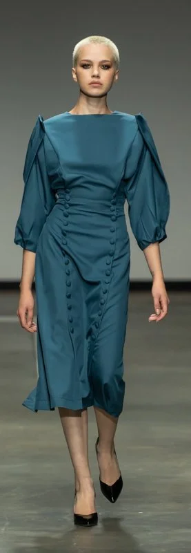 Model wearing a teal long-sleeve dress with button details, walking on a runway.