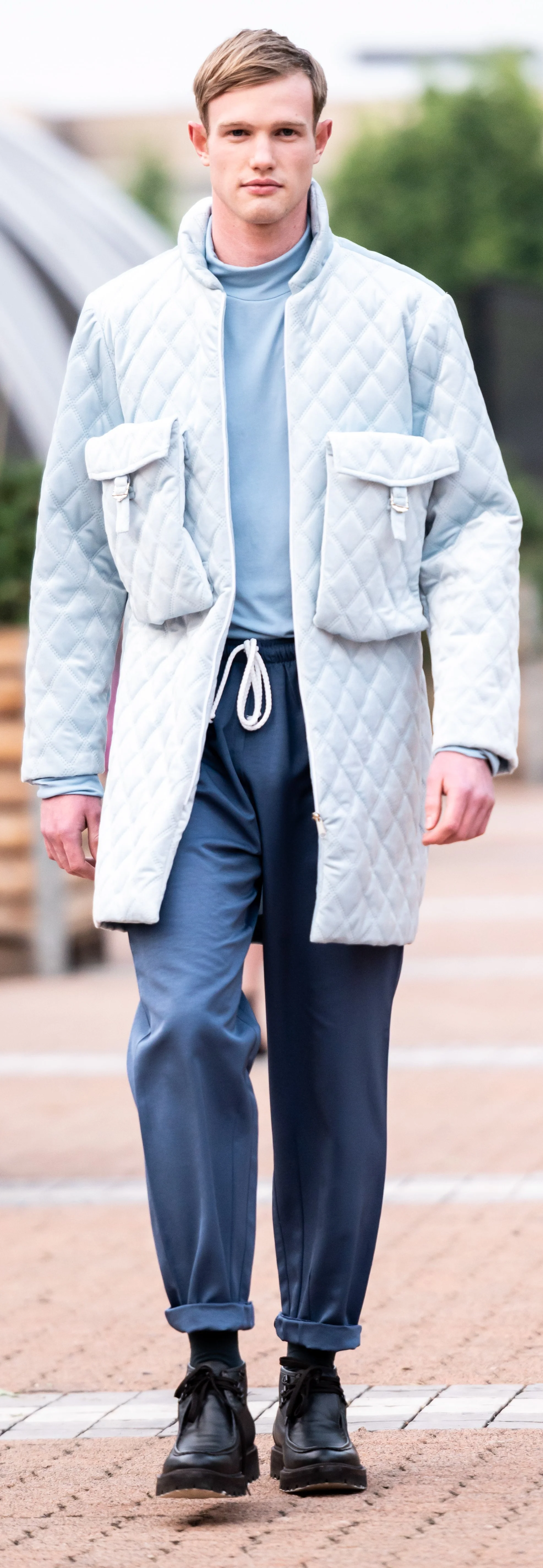 Model wearing a long quilted jacket with large pockets, a blue turtleneck, blue pants with drawstring, and black shoes, walking on a pathway outdoors.
