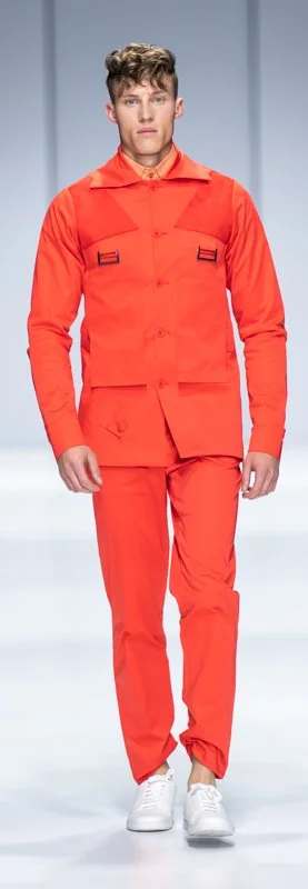Model wearing bright orange outfit with matching pants and jacket, walking on a runway with white sneakers, in a fashion show setting.