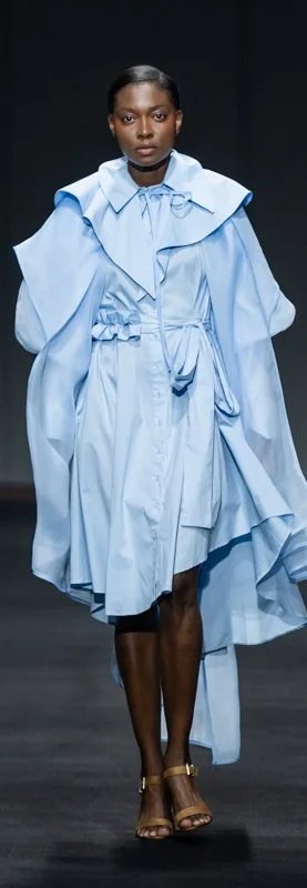 Model wearing a light blue designer dress with layered ruffles and cape-like details, strappy brown sandals, walking on a runway.