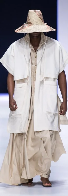 Person wearing a large straw hat, long white coat, and beige flowing outfit on a runway.