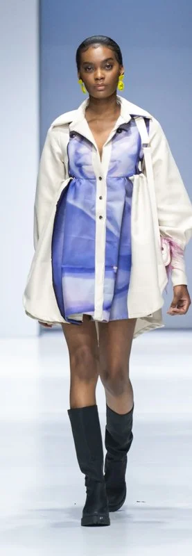 Fashion model on runway wearing a blue dress with a white coat, black boots, and yellow earrings.
