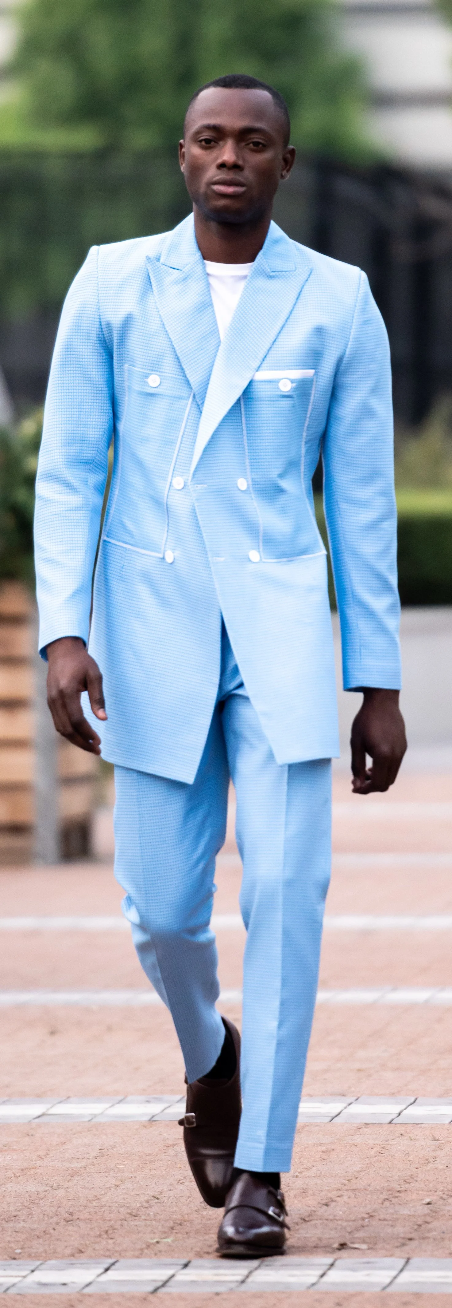 Man walking in light blue double-breasted suit, white shirt, and brown dress shoes outdoors.