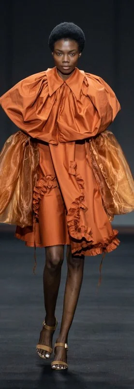 Model on runway wearing an orange dress with ruffles and puffed sleeves