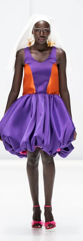Model wearing a purple and orange dress with white headscarf on runway.