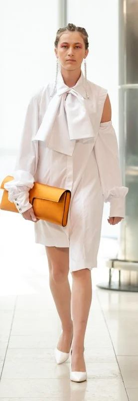 Person in white outfit with large bow and cut-out shoulders, holding an orange clutch, wearing white heels.