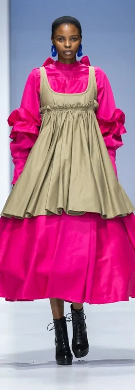 Model in bright pink dress with tan overlay and black boots walking on runway
