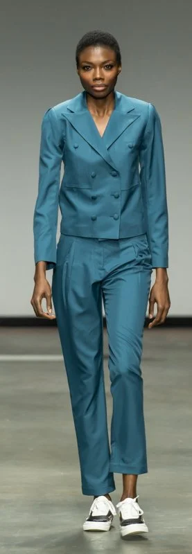 Model walking on runway wearing teal suit with double-breasted jacket and matching pants, paired with white shoes.