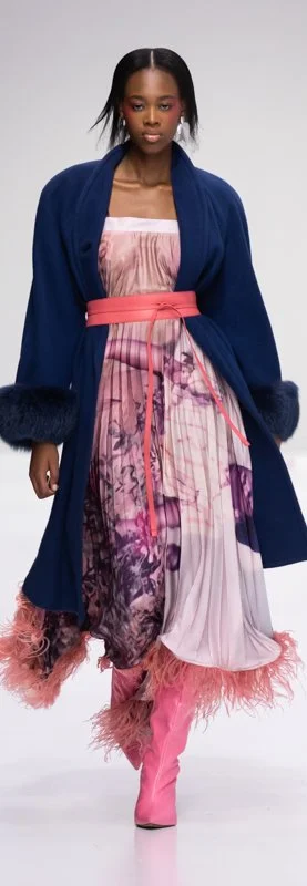 Model wearing a blue coat, pink patterned dress, and pink boots on the runway.