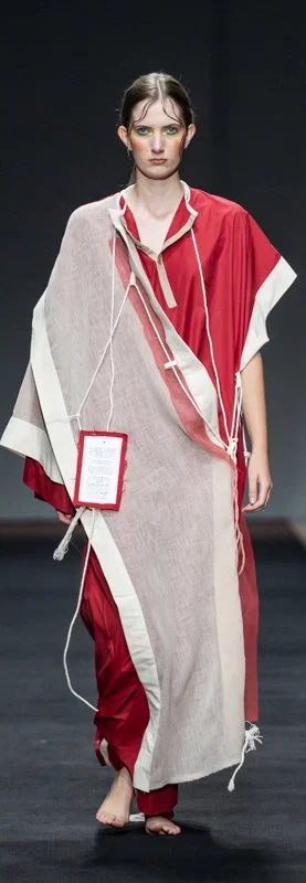 Model wearing a red and beige asymmetrical dress with a square tag on a runway.