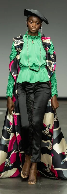 Model on runway wearing a colorful fashion outfit with a green ruffled top, patterned cape, black pants, and a beret.