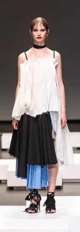 Model walking on runway wearing a layered black, white, and blue dress, with strappy black heels, and a choker necklace.