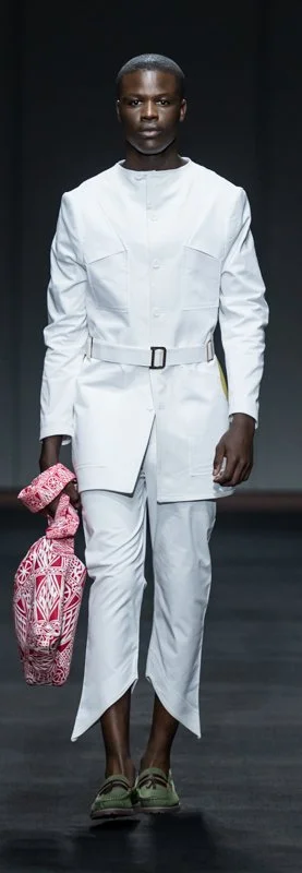 Model walking on runway wearing white outfit with belt and holding red patterned bag