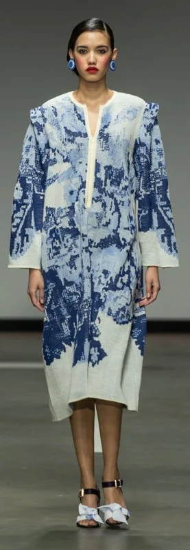 Model wearing a blue and white patterned dress with long sleeves and shoulder design, accessorized with blue earrings and decorative heels on a runway.