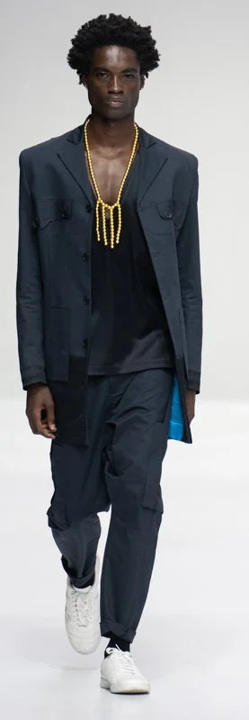 Model wearing dark suit with gold necklace and white sneakers on runway
