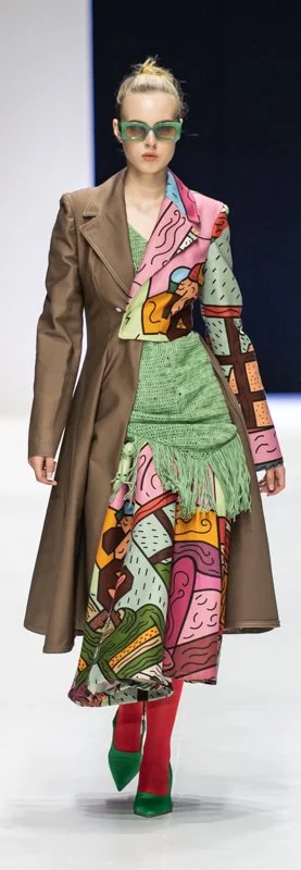 Model wearing a long brown coat with colorful abstract patterns, green knit dress with fringe, red tights, green shoes, and green sunglasses.