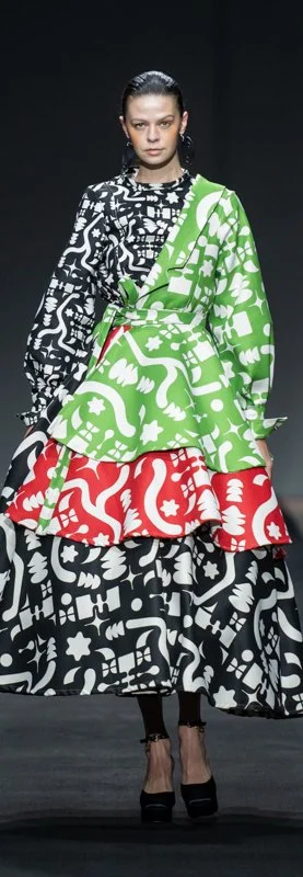 Model wearing a layered dress with black, green, and red abstract patterns on a runway.
