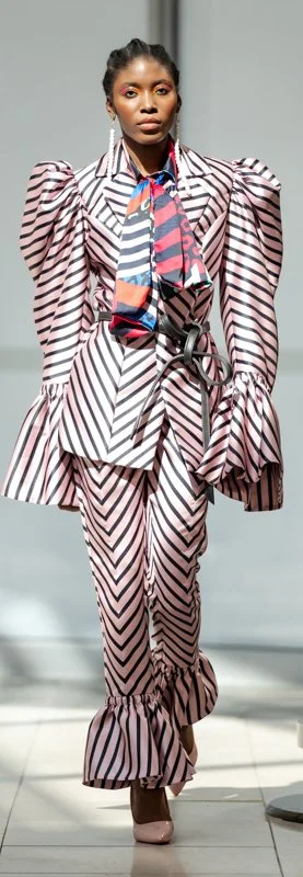 Model in bold striped suit with puffed sleeves, ruffled cuffs, and colorful scarf on runway.