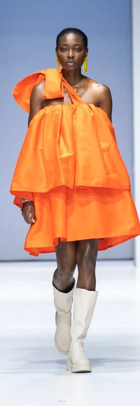 Model on runway wearing an oversized orange dress, yellow earrings, and white boots.