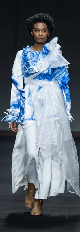 Model wearing a blue and white abstract-patterned outfit with ruffled details, walking on a fashion runway.