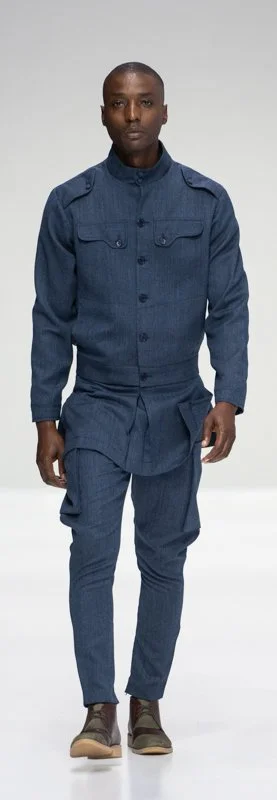 Model walking in a fashion show wearing a blue suit with a military-style jacket and fitted pants, paired with brown shoes.