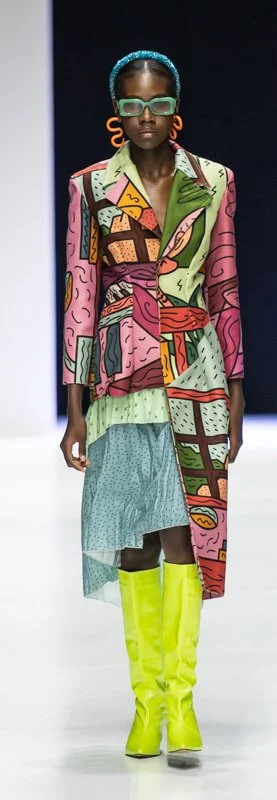 Model wearing a colorful, patterned coat with green abstract designs, neon green boots, orange earrings, and teal sunglasses, walking on a fashion runway.