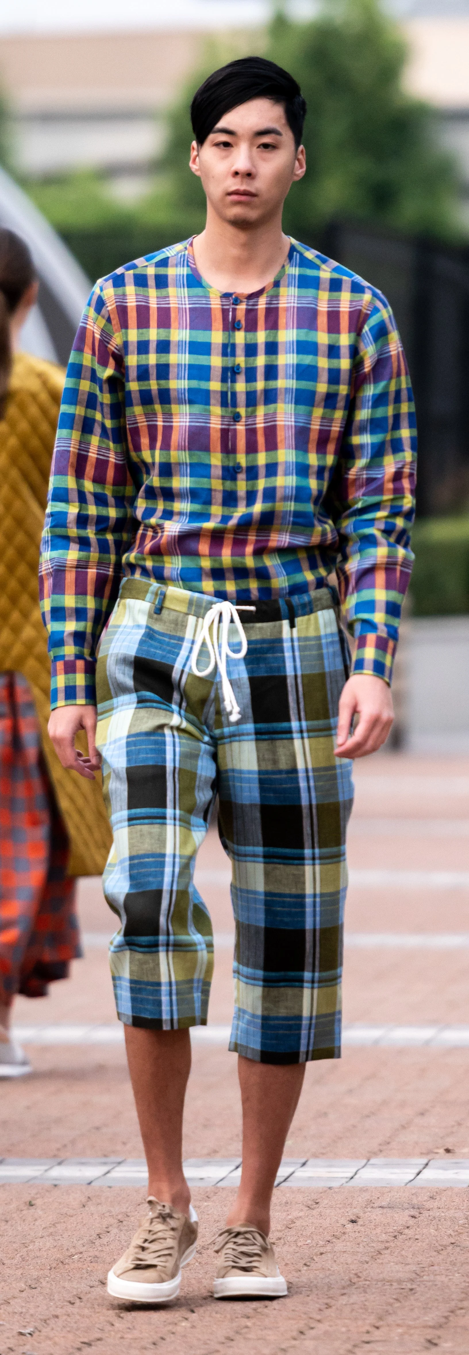 Man wearing a colorful plaid outfit, consisting of a long-sleeve shirt and matching shorts, with tan sneakers, walking outdoors.