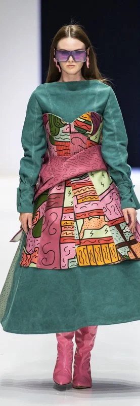 Fashion model wearing a colorful, artistic dress with teal sleeves and abstract patterns, pink boots, purple sunglasses, and matching earrings on a runway.