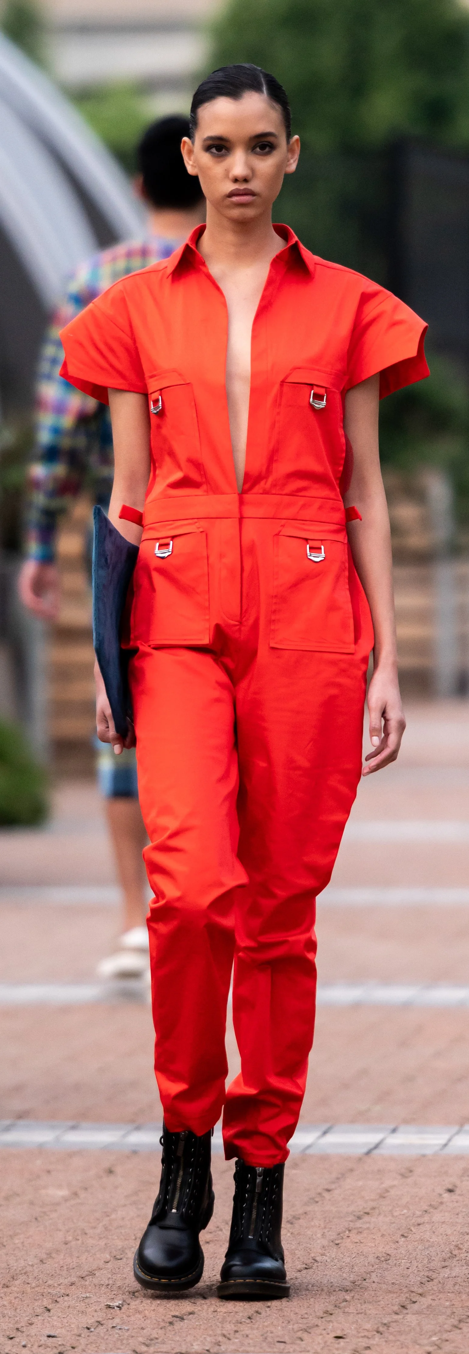Person wearing a bright red jumpsuit with deep neckline and black boots, walking on a pathway, holding a dark clutch. Background is blurred with greenery and another person in casual attire.