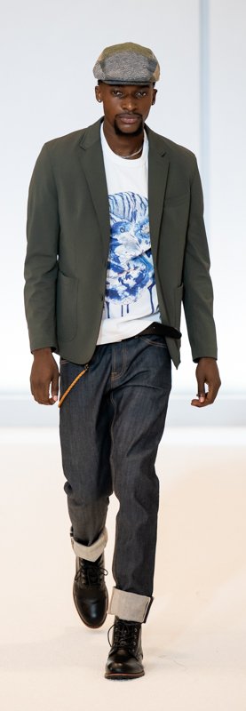 Male model wearing a casual outfit including a gray blazer, graphic t-shirt, rolled-up jeans, and a flat cap, walking on a runway.