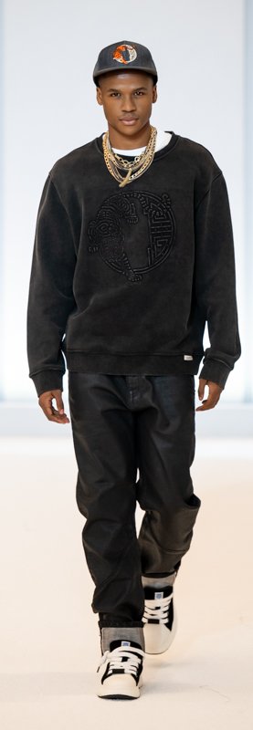 Man walking on runway wearing black sweatshirt, leather pants, sneakers, cap, and gold chains.