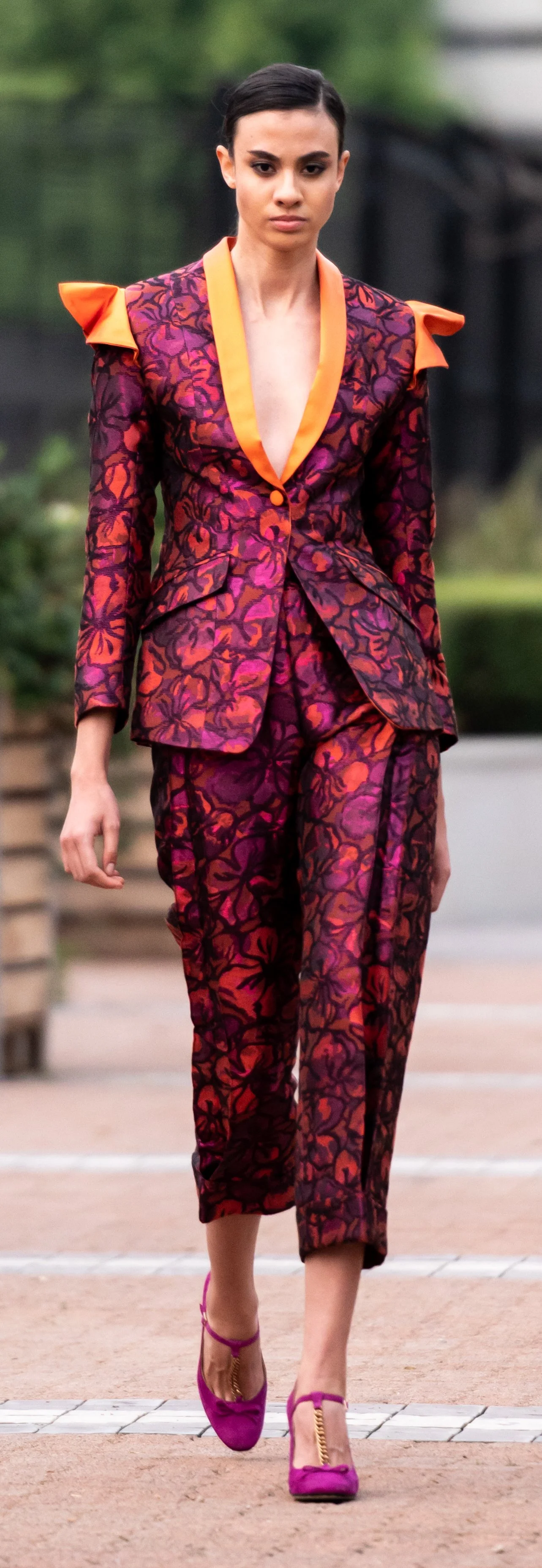 Model wearing a colorful patterned suit with orange lapels and shoulder accents, walking on a runway. The outfit includes matching pants and is paired with magenta shoes.
