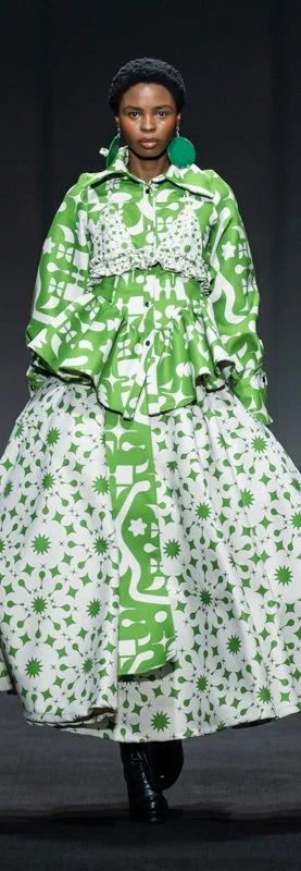 Person wearing a green and white patterned outfit with a floor-length skirt and matching jacket, featuring floral and geometric designs, walking on a runway.