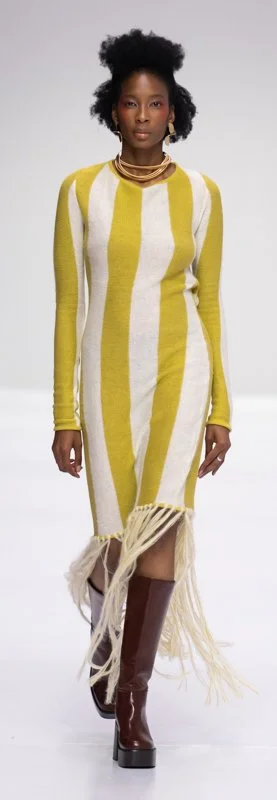 Model wearing a yellow and white striped dress with fringes, brown boots on a runway.