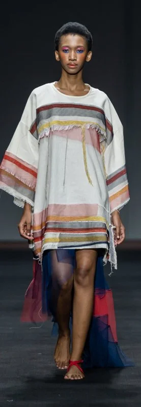 Model walking down runway wearing a colorful, striped kaftan-style dress with frayed edges and layered fabric accents.