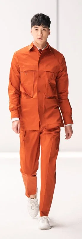 Model wearing an orange outfit with a button-up shirt and matching pants, walking on a runway, paired with white shoes.