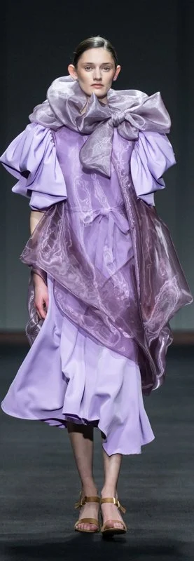 Model wearing a lavender layered dress with ruffled sleeves and large bow on runway