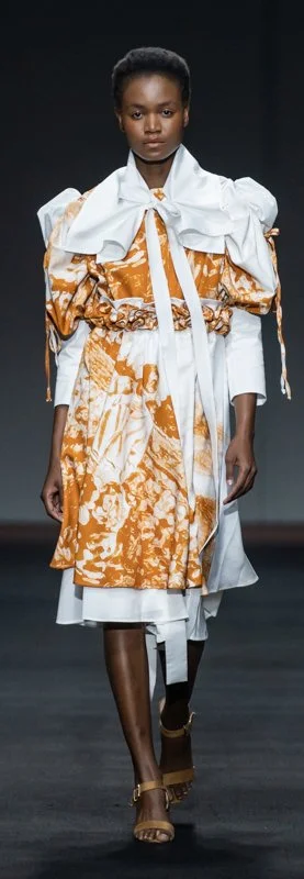 Model walking on runway wearing an elaborate orange and white dress with puffed sleeves and wide belt, beige sandals, fashion show.