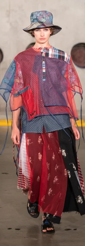 A model walks the runway wearing an eclectic outfit consisting of a colorful patchwork poncho, a wide-brimmed hat, and a long, layered skirt with mixed patterns. The ensemble features reds, blues, and various textures.