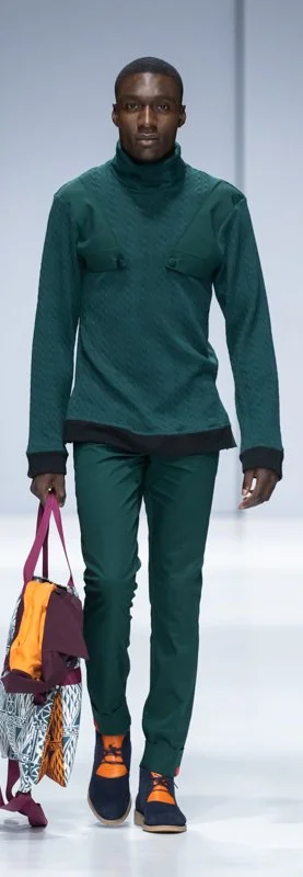 Man walking on runway in green quilted outfit, holding colorful bags