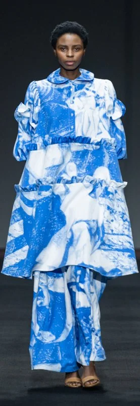 Model wearing a blue and white patterned dress with frills and long sleeves on a runway.